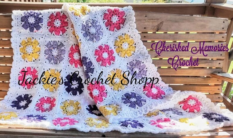May include: A white crochet baby blanket with a scalloped edge. The blanket is made up of squares with different colored flowers in the center. The colors of the flowers are pink, yellow, purple, and white. The text "Jackies Crochet Shopp" and "Cherished Memories Crochet" is visible on the blanket.