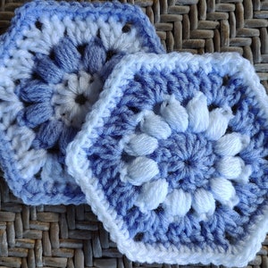 Peut inclure: Deux dessous de verre faits à la main au crochet, dans des tons de bleu et de blanc. Les dessous de verre ont une forme hexagonale avec un motif floral au centre. La laine semble douce et les dessous de verre sont posés sur une surface texturée.