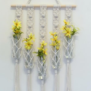 May include: A macrame wall hanging with four glass vases, each holding a bouquet of yellow flowers. The wall hanging has a wooden dowel at the top and is made of white cotton cord.