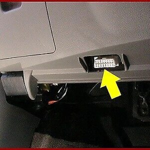 May include: A close-up of a gray car interior with a black plastic connector exposed. The connector is located in a small compartment under the dashboard. A yellow arrow points to the connector.