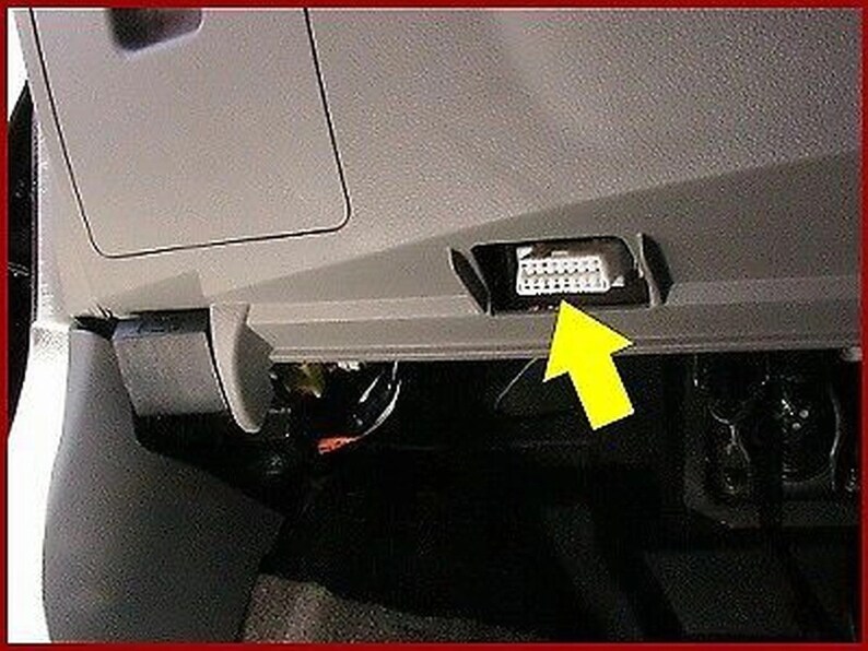 May include: A close-up of a gray car interior with a black plastic connector exposed. The connector is located in a small compartment under the dashboard. A yellow arrow points to the connector.