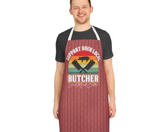 Op de afbeelding: Een rode schort met een herhalend patroon en de tekst "Support Your Local Butcher". De schort heeft een afbeelding van een varken, gekruiste vleesmessen en een zonsondergangontwerp. De schort wordt door een persoon gedragen.
