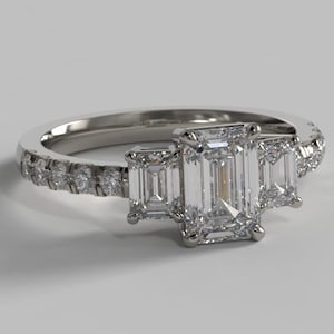 Könnte beinhalten: Ein silberner Verlobungsring mit einem zentralen Smaragdschliff-Diamanten und zwei kleineren Smaragdschliff-Diamanten auf jeder Seite. Die Ringschiene ist mit kleineren runden Diamanten besetzt.