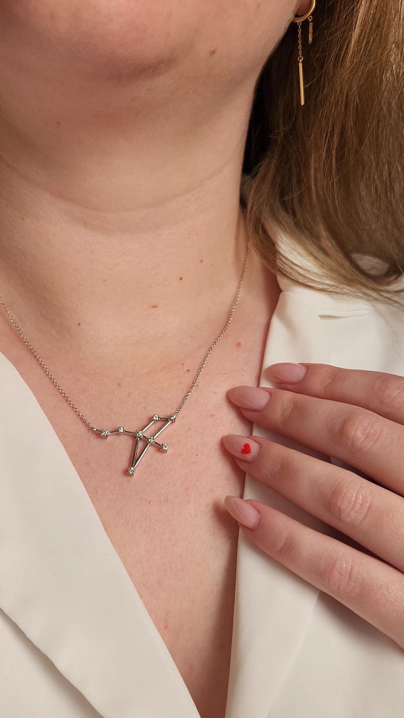 May include: A silver necklace with a constellation pendant featuring small diamonds. The necklace is worn by a person with a white shirt and red nail polish.