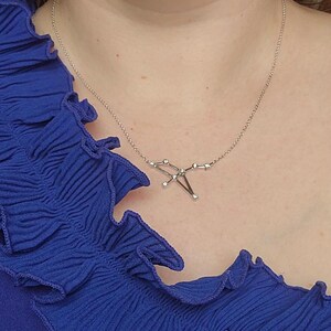 May include: A silver necklace with a constellation design featuring small clear stones. The necklace is worn by a person wearing a blue ruffled top.
