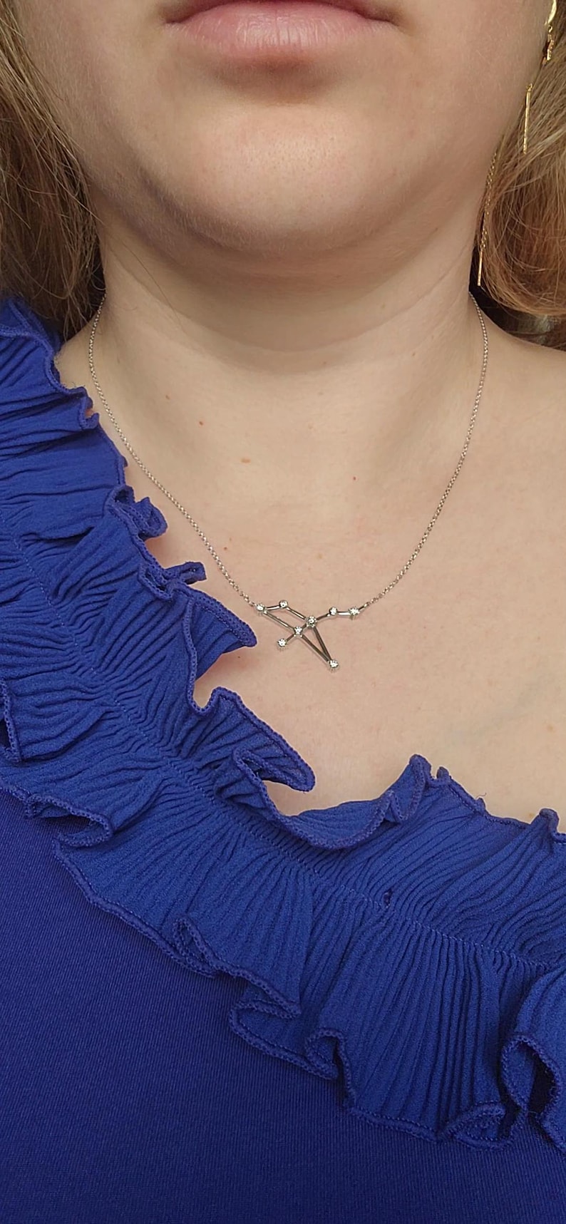 May include: A silver necklace with a constellation design featuring small clear stones. The necklace is worn by a person wearing a blue ruffled top.
