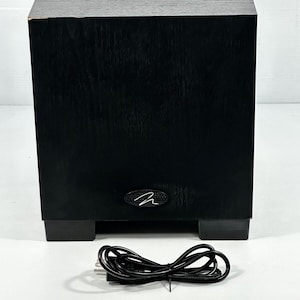 May include: A black, rectangular subwoofer with a wood grain finish. The subwoofer has a small, oval-shaped logo on the front and sits on four black feet. A black power cord is in front of the subwoofer.