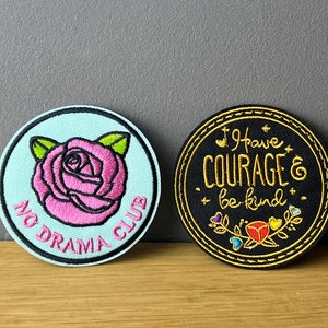 Könnte beinhalten: Zwei gestickte Bügelbilder. Eines ist hellblau mit einer rosa Rose und dem Text "No Drama Club". Das andere ist schwarz mit goldfarbenem Text, der "I have courage & be kind" lautet, und einer kleinen roten Rose.