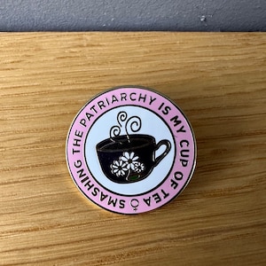 Könnte beinhalten: Runder Emaille-Anstecker mit rosa Rand und goldfarbener Umrandung. Der Anstecker zeigt eine schwarze Teetasse mit weißen Gänseblümchen-Details und Dampf. Der Text "SMASHING THE PATRIARCHY IS MY CUP OF TEA" steht am Rand.