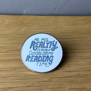 Könnte beinhalten: Eine weiße Emaille-Anstecknadel mit einer blauen Aufschrift, die lautet: "All this reality is really cutting into my reading time".