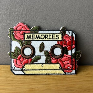 Könnte beinhalten: Weißer gestickter Kassetten-Patch mit roten und grünen Blumendetails. Das Band-Label lautet "MEMORIES".