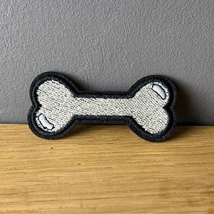 Könnte beinhalten: Gestickter Aufnäher in Form eines Hundeknochens. Der Knochen ist weiß mit schwarzen Konturen und Akzenten. Der Aufnäher ist wahrscheinlich für Kleidung oder Accessoires.