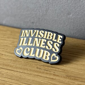 May include: A black pin with the words "INVISIBLE ILLNESS CLUB" in a cream-colored, retro font. The pin has a black border and two cream-colored hearts. The pin is on a wooden surface.