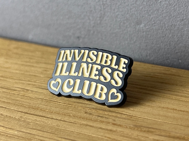 May include: A black pin with the words "INVISIBLE ILLNESS CLUB" in a cream-colored, retro font. The pin has a black border and two cream-colored hearts. The pin is on a wooden surface.