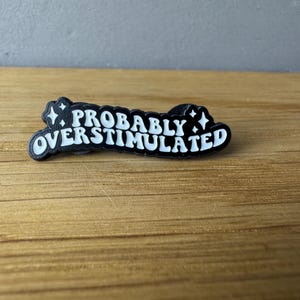 May include: A black and white pin with the phrase "PROBABLY OVERSTIMULATED" in a stylized font. The pin has a black border with white text and star accents. The pin is on a wooden surface.