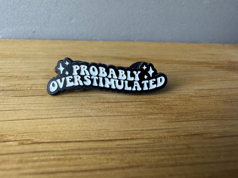 May include: A black and white pin with the phrase "PROBABLY OVERSTIMULATED" in a stylized font. The pin has a black border with white text and star accents. The pin is on a wooden surface.