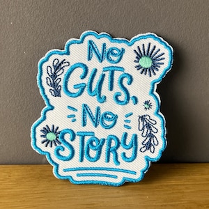 Könnte beinhalten: Gestickter Aufnäher mit dem Schriftzug "No Guts, No Story" in türkisfarbener Schrift. Der Aufnäher hat einen weißen Hintergrund mit türkisfarbenem Rand und dekorativen floralen Elementen.