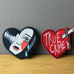 Könnte beinhalten: Zwei gestickte Bügelbilder in Herzform. Das erste Patch ist schwarz mit einem weißen Gesicht einer Frau mit einem blauen Auge und einer roten Wange. Die Frau weint blaue Tränen. Das zweite Patch ist rot mit einem weißen Messer und den Worten "TRUE CRIME" in weiß.