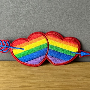 Könnte beinhalten: Gestickter Aufnäher mit zwei Herzen in Regenbogenfarben, durchbohrt von einem blauen Pfeil. Die Herzen haben einen roten Rand. Der Aufnäher ist für Kleidung oder Accessoires.
