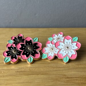 Könnte beinhalten: Zwei Emaille-Pins mit rosa und weißen Blumen mit goldenen Akzenten. Die Blumen sind in einer Gruppe angeordnet und haben einen 3D-Effekt. Ein Pin zeigt schwarze Blumen mit rosa Akzenten, während der andere weiße Blumen mit rosa Akzenten zeigt.