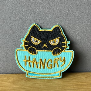 Könnte beinhalten: Eine schwarze Katze mit wütenden Augen schaut über den Rand einer hellblauen Schüssel mit dem in goldenen Fäden gestickten Wort "HANGRY".