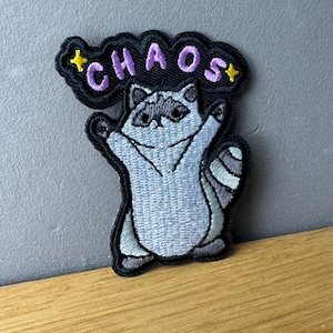 Könnte beinhalten: Gestickter Aufnäher mit einem Cartoon-Waschbären, der die Arme hebt. Der Aufnäher hat einen schwarzen Rand und das Wort "CHAOS" in lila Schrift über dem Waschbären, mit gelben Sternen auf beiden Seiten. Der Waschbär ist hauptsächlich grau und weiß.