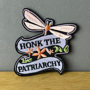 Könnte beinhalten: Gestickter Aufnäher mit einem Libellen-Design und den Worten "HONK THE PATRIARCHY" in schwarzer Schrift auf einem hellblauen Banner. Der Aufnäher zeigt orangefarbene Blumen und grüne Blätter.