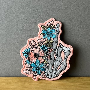 Könnte beinhalten: Ein rosa gestickter Patch mit einer Skeletthand, die einen Strauß rosa, blauer und grüner Blumen hält.