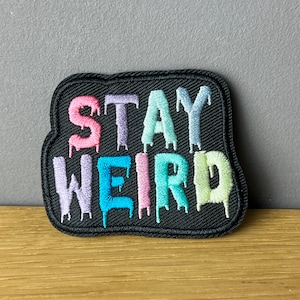 Könnte beinhalten: Ein schwarzer gestickter Aufnäher mit dem Text "STAY WEIRD" in Regenbogenfarben. Die Buchstaben tropfen mit einem tropfenden Farbeffekt.