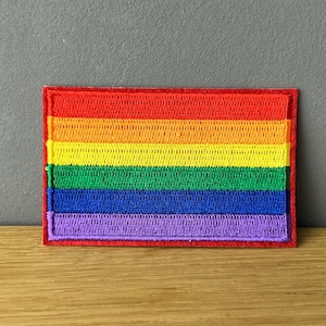 Könnte beinhalten: Ein Regenbogen-Pride-Flaggen-Patch mit roten, orangen, gelben, grünen, blauen und violetten Streifen. Der Patch ist gestickt und hat einen roten Rand.