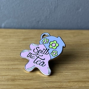 Könnte beinhalten: Emaille-Anstecknadel mit einer hellblauen Teekanne mit grünen und gelben Blumen, die eine hellviolette Flüssigkeit verschüttet. Der Text "Spill the Tea" ist in schwarzer Schrift auf dem violetten Klecks geschrieben. Die Nadel hat einen silbernen Rand.