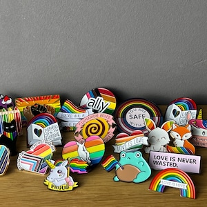 Könnte beinhalten: Eine Sammlung von Emaille-Pins mit Regenbogenfarben und LGBTQ+-Motiven, darunter "Love is Love", "You are Safe with Love", "Love is Never Wasted", "Libraries are for Everyone", "We're All a Little Bit Cinnamon Rolls", "Ally", "Proud" und "I'd Rather".