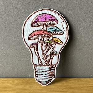 Könnte beinhalten: Gestickter Aufnäher mit einer Gruppe bunter Pilze in einer Glühbirnenform. Die Pilze haben rosa, blaue, rote, gelbe und violette Kappen. Der Aufnäher hat einen weißen Hintergrund mit braunen Umrissen.