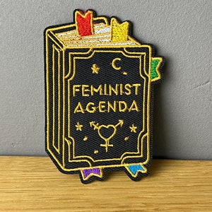 Könnte beinhalten: Schwarzer, gestickter Aufnäher in Form eines Buches mit den Worten "FEMINIST AGENDA". Der Aufnäher hat goldfarbene Verzierungen und bunte Schleifen-Lesezeichen. Ein Symbol eines Herzens mit Kreuz und Pfeil befindet sich in der Mitte des Aufnähers.