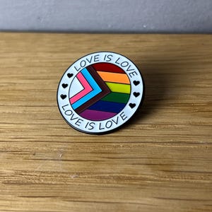 Könnte beinhalten: Ein runder Emaille-Ansteckpin mit weißem Rand und dem schwarzen Schriftzug "LOVE IS LOVE". Der Pin zeigt ein Regenbogen-Design mit rosa, blau, braun, orange, gelb, grün, lila und weinroten Farben. Kleine schwarze Herzen umranden den Rand.