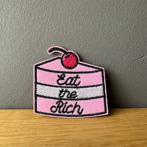 Könnte beinhalten: Gestickter Aufnäher in Form eines rosa Kuchenstücks mit einer Kirsche oben. Der Text "Eat the Rich" ist in schwarzer Schrift über den Kuchen gestickt. Der Aufnäher hat einen schwarzen Rand.