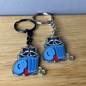 Könnte beinhalten: Zwei Schlüsselanhänger aus Acryl mit einem Cartoon-Waschbären, der aus einer blauen Mülltonne lugt. Die Schlüsselanhänger haben einen silbernen und schwarzen Metallring und eine Kette. Die Mülltonne hat einen blauen Deckel und rote Stiefel.
