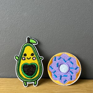 Könnte beinhalten: Zwei gestickte Bügelbilder, eines mit einem lächelnden Avocado mit einem Herzen in der Mitte und das andere mit einem lila Donut mit blauen Streuseln.