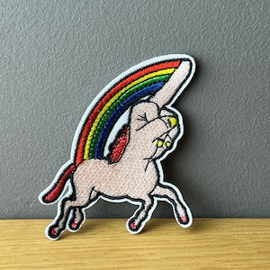 Könnte beinhalten: Gestickter Aufnäher mit einem hellrosa Einhorn mit Regenbogenmähne und -schwanz. Das Einhorn ist in Bewegung, mit einem Regenbogen über seinem Rücken. Der Aufnäher hat einen weißen Rand.