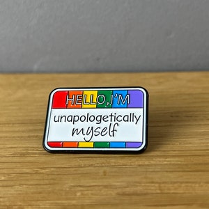 Könnte beinhalten: Eine Regenbogen-Emaille-Anstecknadel mit schwarzem Rand. Die Anstecknadel hat die Form eines Namensschildes und lautet "HELLO, I'M unapologetically myself".