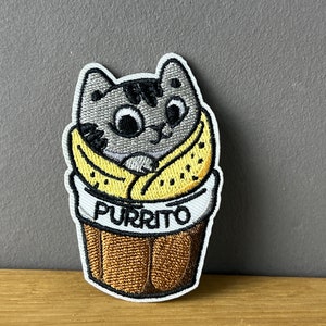 Könnte beinhalten: Gestickter Aufnäher mit einer grauen Katze, die in eine gelbe Tortilla gewickelt ist, wie ein Burrito. Der Aufnäher trägt den Schriftzug "PURRITO" in weißen Buchstaben. Die Katze hat große Augen und einen niedlichen Ausdruck.