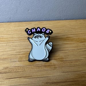 Könnte beinhalten: Ein hellgrauer Emaille-Pin mit einem Waschbären und dem Wort "CHAOS" in lila Buchstaben darüber. Der Waschbär hat die Arme erhoben und ist schwarz umrandet. Der Pin befindet sich auf einer Holzoberfläche.