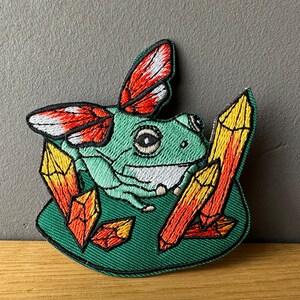Könnte beinhalten: Gestickter Bügel-Patch mit einem grünen Frosch mit roten und weißen Schmetterlingsflügeln, umgeben von orangefarbenen und gelben Kristallen auf grünem Hintergrund.