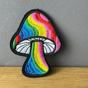Könnte beinhalten: Ein gestickter Bügel-Patch in Form eines Regenbogenpilzes. Der Pilz hat einen weißen Hut mit schwarzem Rand und einen regenbogenfarbenen Stiel.