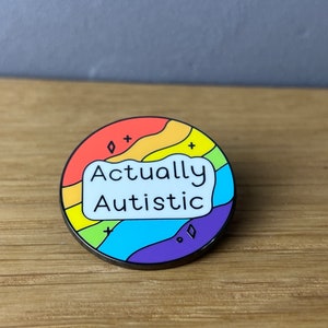 Könnte beinhalten: Runder Emaille-Pin mit Regenbogen-Design und den Worten "Actually Autistic" in einer weißen Sprechblase. Der Pin hat einen schwarzen Rand und ist vor einem hellgrauen Hintergrund platziert.