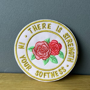 Könnte beinhalten: Ein weißer gestickter Aufnäher mit einem goldenen Kreis um den Rand. Der Text im Kreis lautet "There is strength in your softness." Zwei rosa Rosen sind in der Mitte des Aufnähers gestickt.