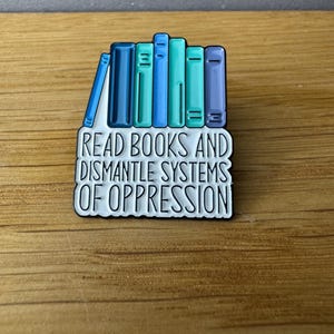 Könnte beinhalten: Anstecknadel mit einem Design aus gestapelten Büchern in Blau- und Grüntönen. Der Text "READ BOOKS AND DISMANTLE SYSTEMS OF OPPRESSION" ist in Schwarz und Weiß dargestellt. Die Nadel befindet sich auf einem Holzuntergrund.