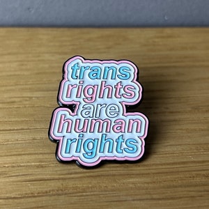 Könnte beinhalten: Eine rosa und blaue Emaille-Anstecknadel mit dem Text "trans rights are human rights".