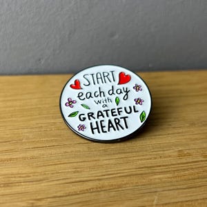 Könnte beinhalten: Weißer Emaille-Pin mit schwarzem Rand. Der Pin zeigt ein rotes Herz, rosa Blumen, grüne Blätter und den Text "START each day with a GRATEFUL HEART".