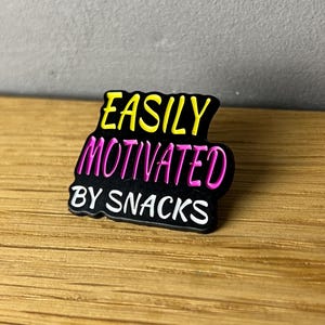 Könnte beinhalten: Schwarzer Anstecker mit dem Text "EASILY MOTIVATED BY SNACKS". Der Text ist in Gelb, Pink und Weiß gehalten. Der Anstecker liegt auf einer Holzoberfläche.